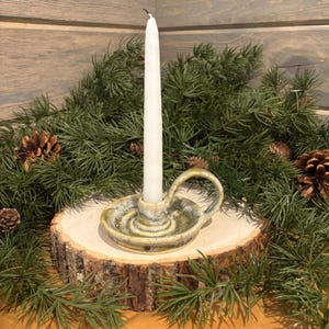 May include: A ceramic candle holder with a white candle. The holder is glazed in shades of green and tan, with a small handle. The candle holder sits on a wooden slice, surrounded by pine branches and pine cones.