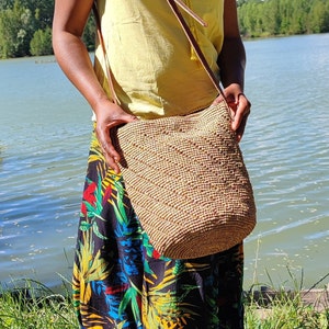 May include: A woven beige shoulder bag with a brown leather strap. The bag is cylindrical and has a textured surface. The person is wearing a yellow top and a long skirt with a colorful floral pattern.