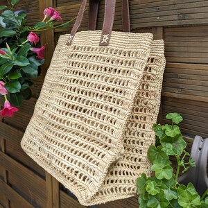 K&ouml;nnte beinhalten: Eine beige geflochtene Strohtasche mit braunen Ledergriffen. Die Tasche h&auml;ngt an einem Holzzaun.
