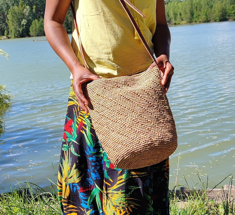 May include: A woven beige shoulder bag with a brown leather strap. The bag is cylindrical and has a textured surface. The person is wearing a yellow top and a long skirt with a colorful floral pattern.