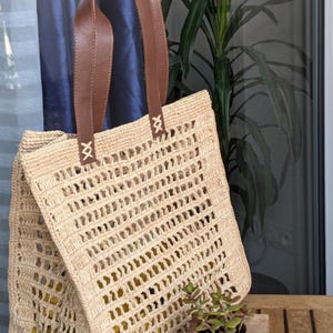 K&ouml;nnte beinhalten: Eine beige geh&auml;kelte Tragetasche mit braunen Ledergriffen. Die Tasche hat ein gewebtes Muster und ist aus Naturfasern gefertigt.