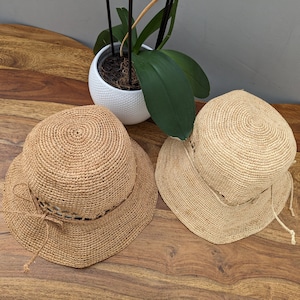 Peut inclure: Deux chapeaux de soleil en paille avec une texture tissée et une garniture de ruban marron. Les chapeaux sont posés sur une surface en bois.