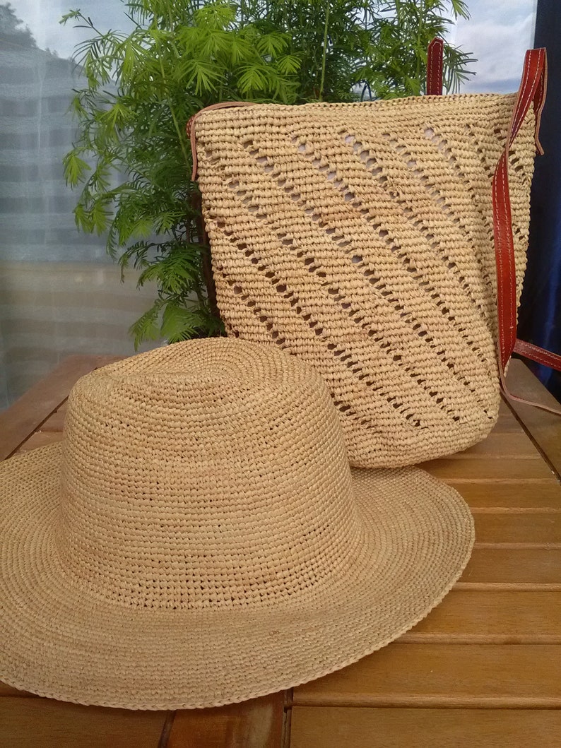 May include: A woven straw hat with a wide brim and a matching backpack with leather straps. The hat is lying on a wooden surface next to the backpack. The backpack has a diagonal pattern.