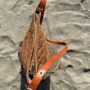 May include: A brown woven shoulder bag with a leather strap and a zipper closure. The bag is made of a textured material and has a rectangular shape. The leather strap is adjustable and has a silver buckle.