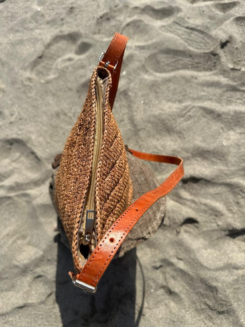 May include: A brown woven shoulder bag with a leather strap and a zipper closure. The bag is made of a textured material and has a rectangular shape. The leather strap is adjustable and has a silver buckle.