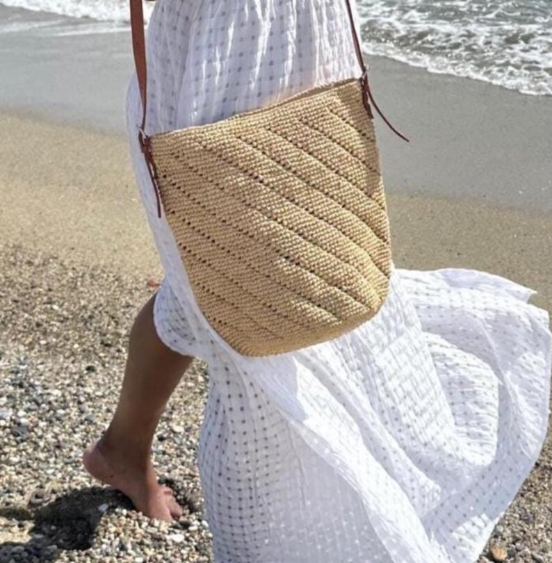 May include: A beige woven shoulder bag with a diagonal pattern and brown leather straps. The bag is worn over a white dress on a beach.