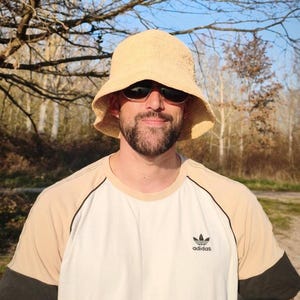 Puede incluir: Una persona con un sombrero de pescador de paja y gafas de sol. La persona lleva una camiseta crema y blanca con detalles negros y el logo de Adidas.