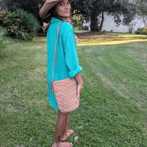 May include: A woman wearing a wide-brimmed straw hat and a turquoise dress. A woven, tan-colored shoulder bag with a diagonal pattern hangs from her shoulder. She is standing on a grassy lawn, with trees in the background.
