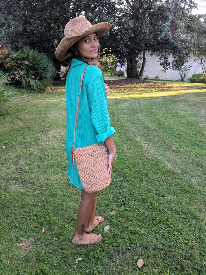 May include: A woman wearing a wide-brimmed straw hat and a turquoise dress. A woven, tan-colored shoulder bag with a diagonal pattern hangs from her shoulder. She is standing on a grassy lawn, with trees in the background.