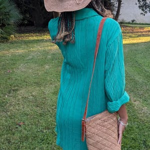 May include: A woman wearing a turquoise green dress, a straw hat with a bow, and a woven shoulder bag with a brown leather strap. The outfit is completed with brown sandals. The background is a grassy lawn.