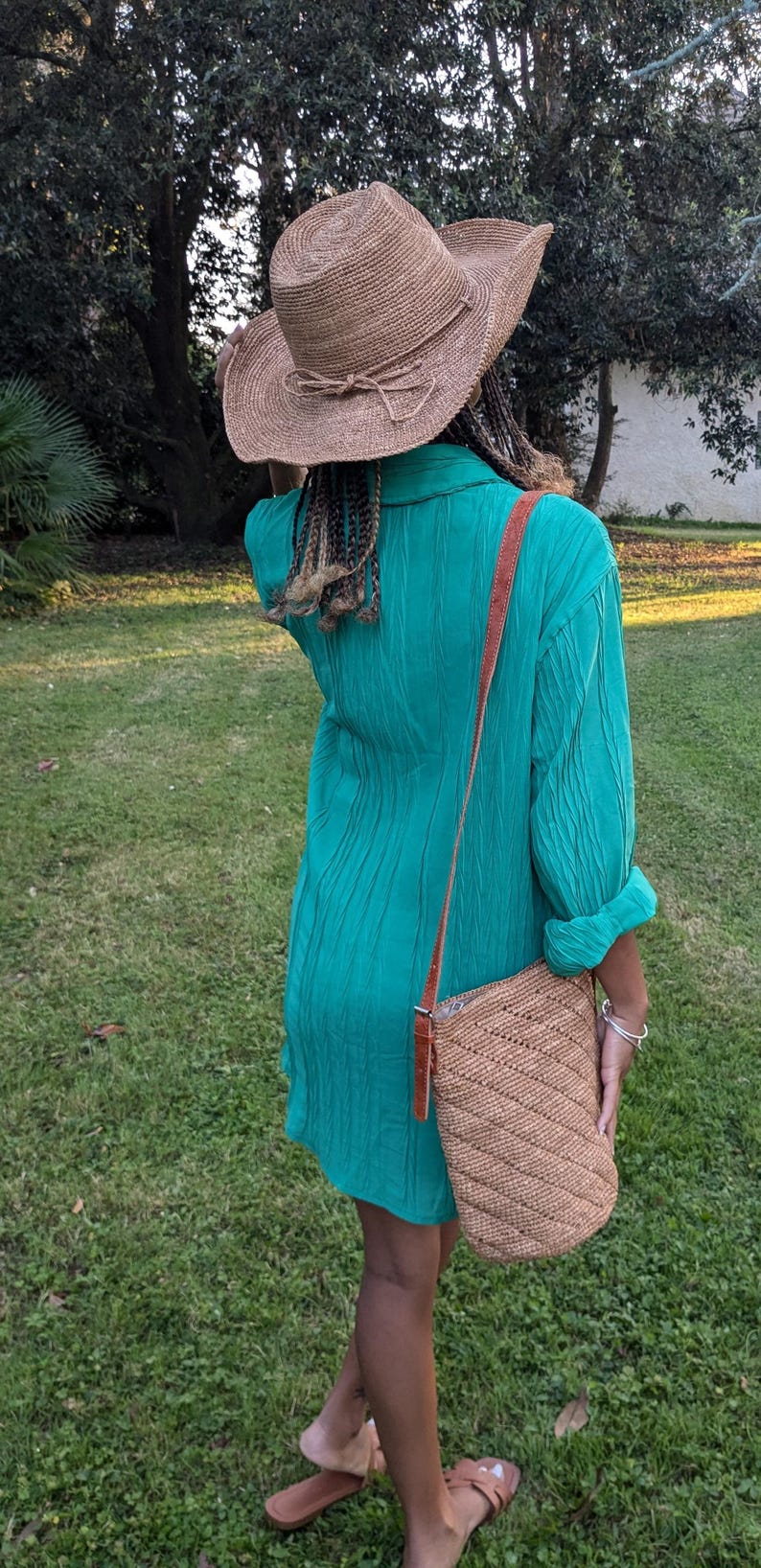 May include: A woman wearing a turquoise green dress, a straw hat with a bow, and a woven shoulder bag with a brown leather strap. The outfit is completed with brown sandals. The background is a grassy lawn.