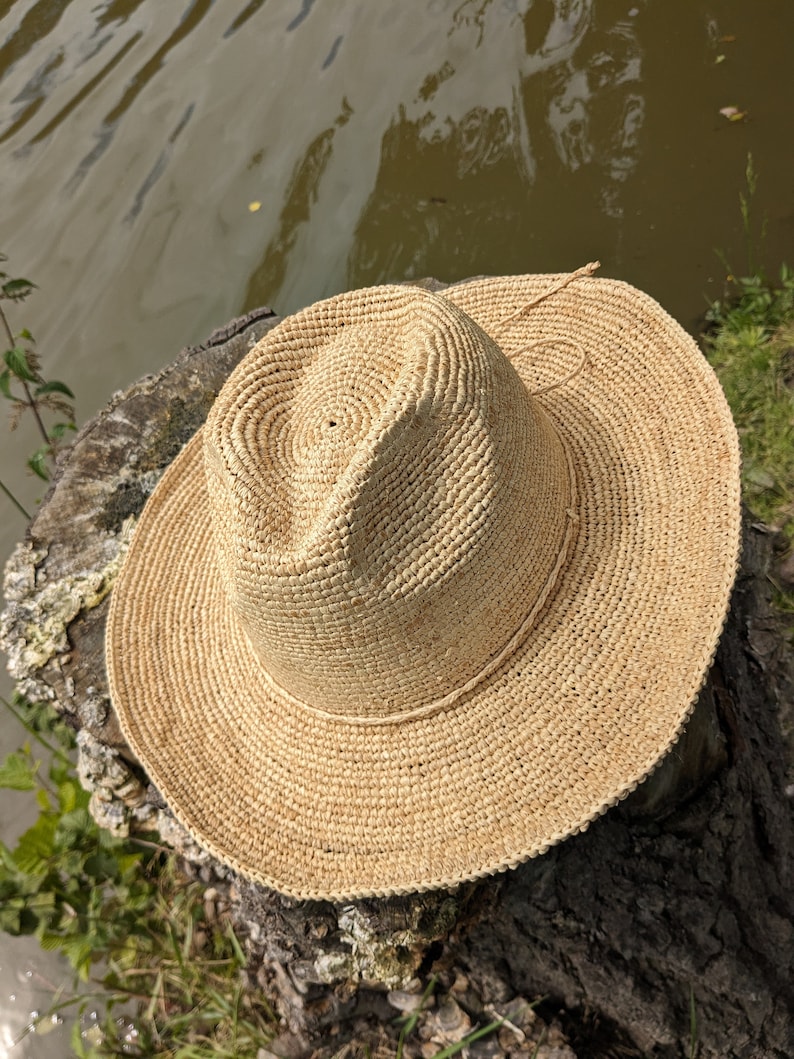 Pode incluir: Um chap&eacute;u de palha castanho claro com uma aba larga e uma faixa estreita em torno da coroa. O chap&eacute;u est&aacute; sentado em um toco de &aacute;rvore.