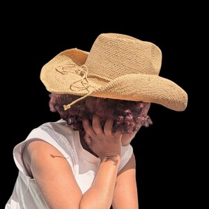 Pode incluir: Uma pessoa usando um chap&eacute;u de cowboy de palha marrom com uma fita marrom. A pessoa est&aacute; usando uma camisa branca sem mangas e tem a cabe&ccedil;a baixa.