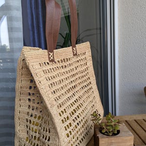 K&ouml;nnte beinhalten: Eine gro&szlig;e, beige geh&auml;kelte Tragetasche mit braunen Ledergriffen. Die Tasche hat ein gewebtes, offenes Design und steht auf einem Holztisch.