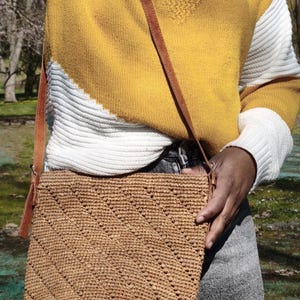 May include: A woven, light brown crossbody bag with a brown leather strap. The bag has a textured, diagonal pattern. The person is wearing a yellow and white sweater and gray jeans.