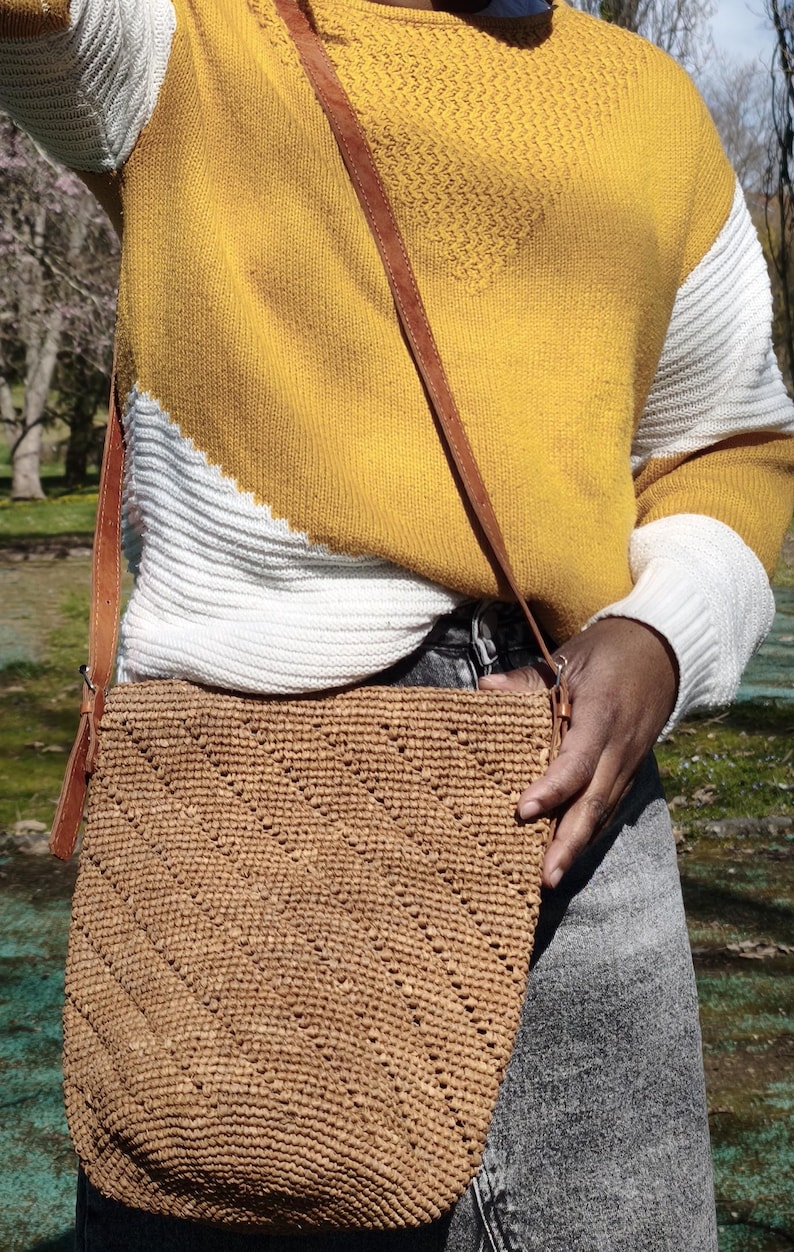 May include: A woven, light brown crossbody bag with a brown leather strap. The bag has a textured, diagonal pattern. The person is wearing a yellow and white sweater and gray jeans.