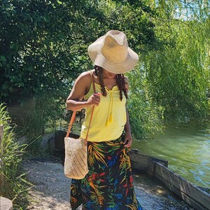 May include: A straw hat, yellow tank top, and a long black skirt with a colorful floral pattern. A woven bag with a brown strap is carried over the shoulder. The person is walking on a gravel path near a lake.