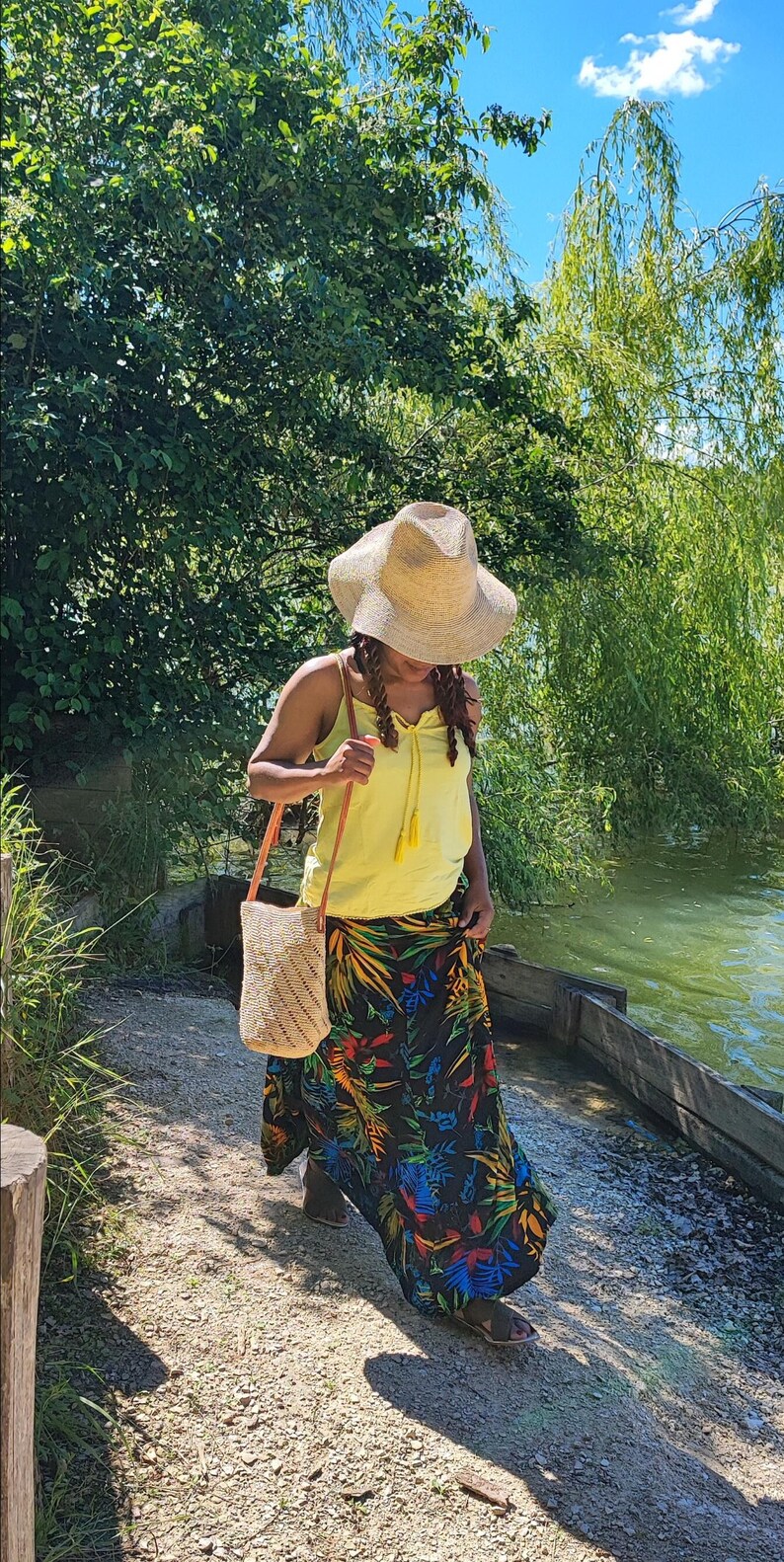 May include: A straw hat, yellow tank top, and a long black skirt with a colorful floral pattern. A woven bag with a brown strap is carried over the shoulder. The person is walking on a gravel path near a lake.