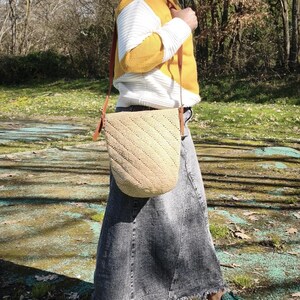 May include: A beige woven shoulder bag with a diagonal pattern and brown leather straps. The bag is carried by a person wearing a yellow and white sweater and a denim skirt. The bag is approximately 12 inches wide and 10 inches tall.