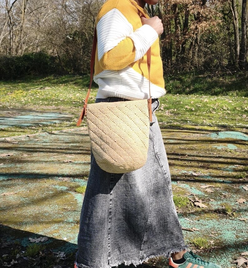 May include: A beige woven shoulder bag with a diagonal pattern and brown leather straps. The bag is carried by a person wearing a yellow and white sweater and a denim skirt. The bag is approximately 12 inches wide and 10 inches tall.
