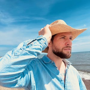 Pode incluir: Um homem vestindo uma camisa de brim azul claro e um chap&eacute;u de cowboy de palha, em p&eacute; em uma praia com o oceano ao fundo.