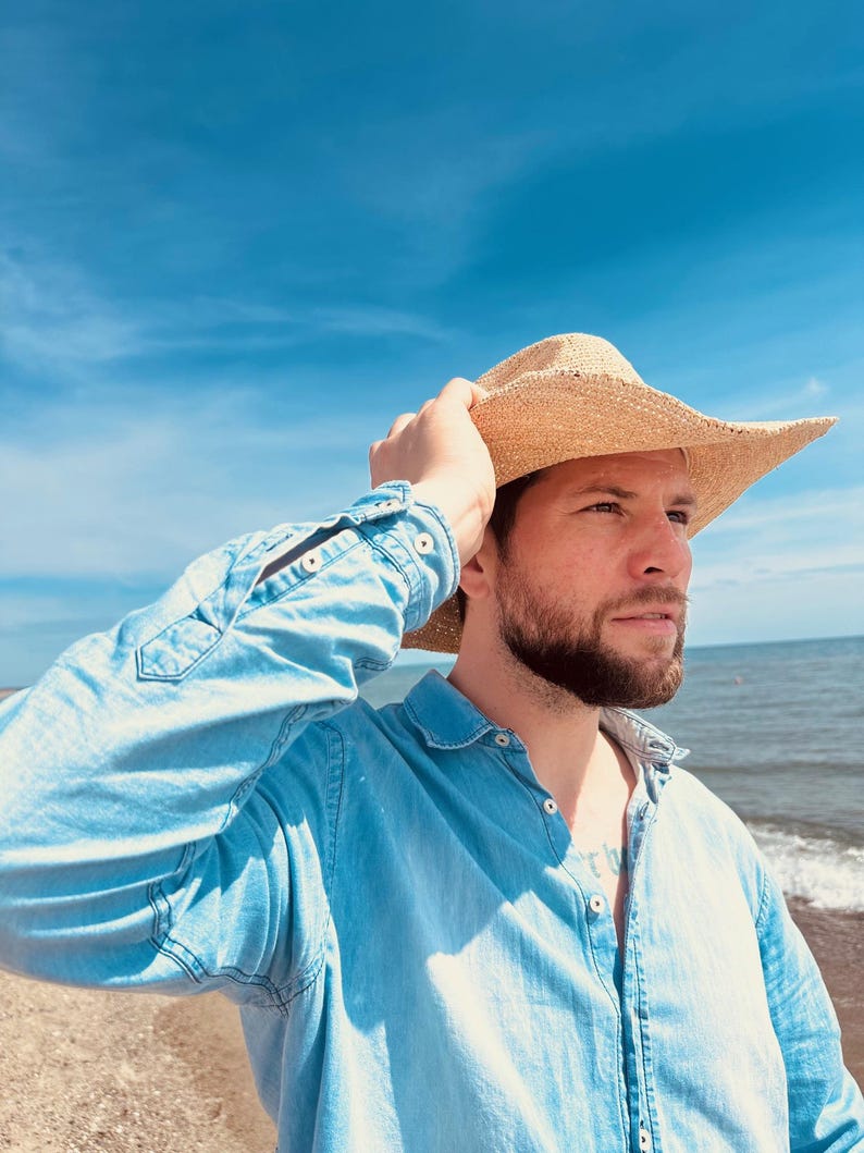 Pode incluir: Um homem vestindo uma camisa de brim azul claro e um chap&eacute;u de cowboy de palha, em p&eacute; em uma praia com o oceano ao fundo.