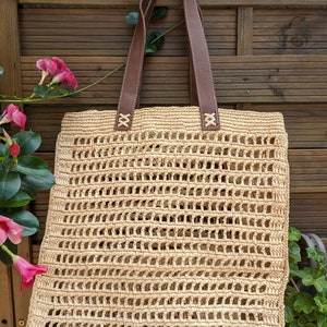 K&ouml;nnte beinhalten: Eine beige geh&auml;kelte Tragetasche mit braunen Lederriemen. Die Tasche h&auml;ngt an einem Holzzaun.