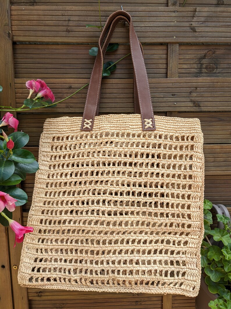 K&ouml;nnte beinhalten: Eine beige geh&auml;kelte Tragetasche mit braunen Lederriemen. Die Tasche h&auml;ngt an einem Holzzaun.