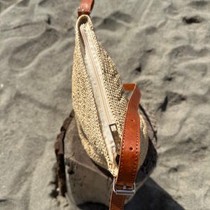 May include: A beige woven handbag with a brown leather strap and a zipper closure. The bag is sitting on a wooden surface, with a sandy background.