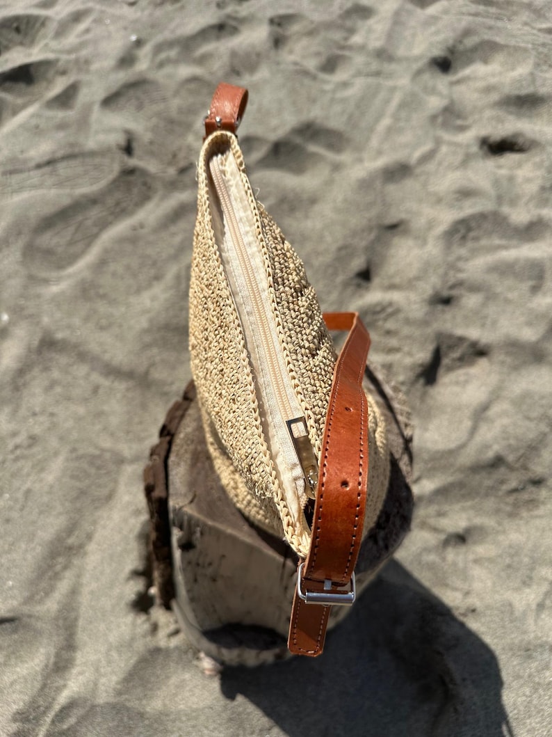 May include: A beige woven handbag with a brown leather strap and a zipper closure. The bag is sitting on a wooden surface, with a sandy background.
