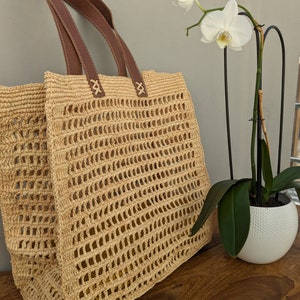 K&ouml;nnte beinhalten: Eine gro&szlig;e, beige, geh&auml;kelte Tragetasche mit braunen Ledergriffen. Die Tasche hat ein gewebtes, offenes Design. Die Tasche steht auf einer Holzoberfl&auml;che neben einer wei&szlig;en Orchideenpflanze in einem wei&szlig;en Topf.
