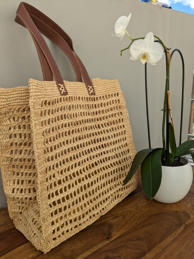 K&ouml;nnte beinhalten: Eine gro&szlig;e, beige, geh&auml;kelte Tragetasche mit braunen Ledergriffen. Die Tasche hat ein gewebtes, offenes Design. Die Tasche steht auf einer Holzoberfl&auml;che neben einer wei&szlig;en Orchideenpflanze in einem wei&szlig;en Topf.