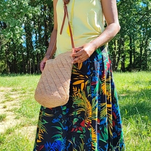 May include: A beige, woven shoulder bag with a diagonal pattern. The bag has a brown leather strap and a yellow tassel. The person is wearing a yellow top and a long black skirt with a colorful floral print.