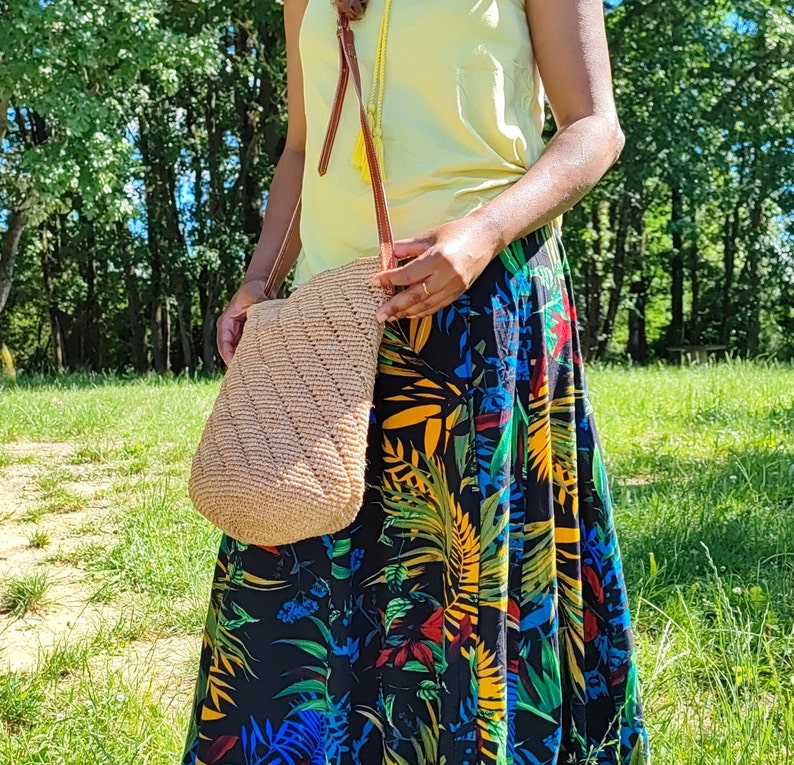 May include: A beige, woven shoulder bag with a diagonal pattern. The bag has a brown leather strap and a yellow tassel. The person is wearing a yellow top and a long black skirt with a colorful floral print.