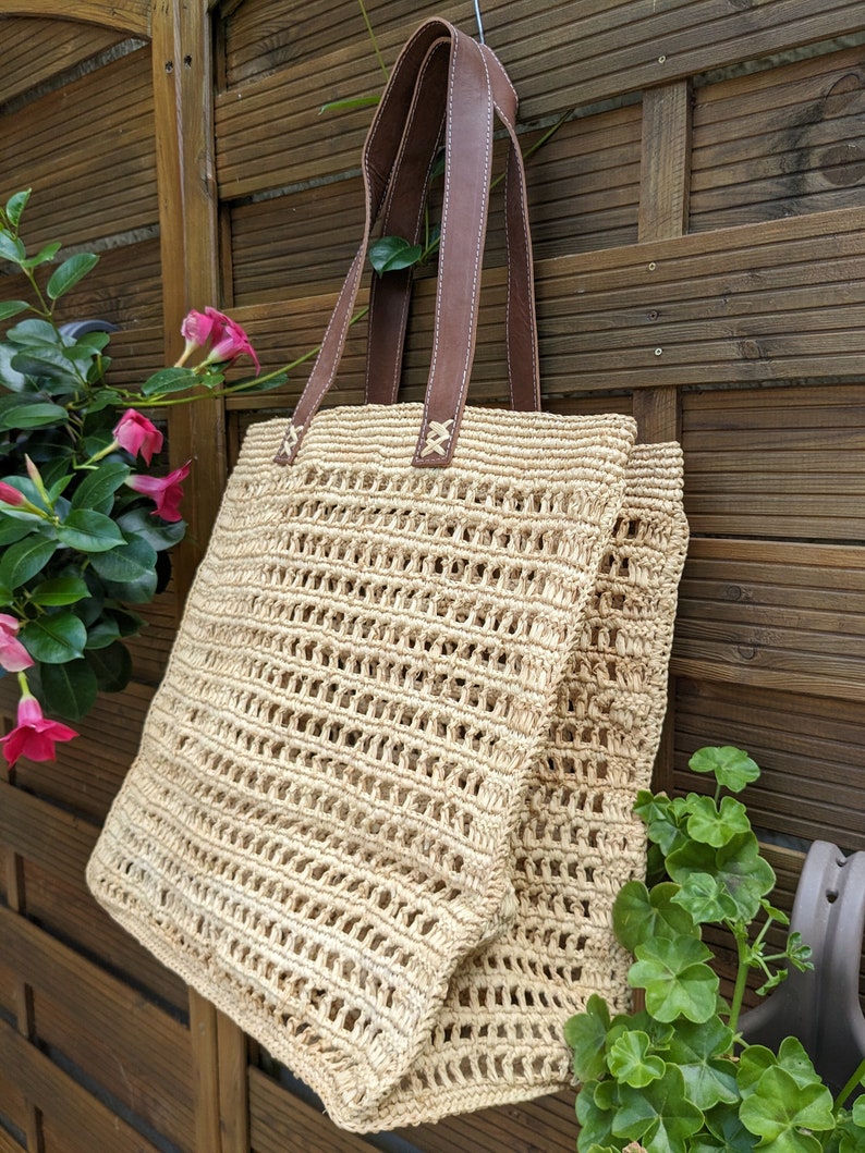 K&ouml;nnte beinhalten: Eine beige geflochtene Strohtasche mit braunen Ledergriffen. Die Tasche h&auml;ngt an einem Holzzaun.