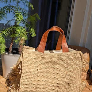 May include: A rectangular woven straw tote bag featuring a natural tan color, two brown leather handles, and decorative fringe detailing along both side edges. The interior is lined with a light-colored fabric, and the bag has a structured shape.