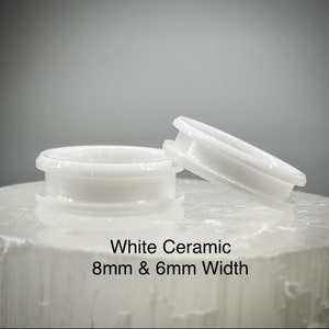 K&ouml;nnte beinhalten: Zwei Ringe aus wei&szlig;er Keramik mit glatter, gl&auml;nzender Oberfl&auml;che. Ein Ring liegt flach, w&auml;hrend der andere schr&auml;g steht. Der Text White Ceramic 8mm & 6mm Width ist unter den Ringen aufgedruckt und gibt das Material sowie die Breiten an.