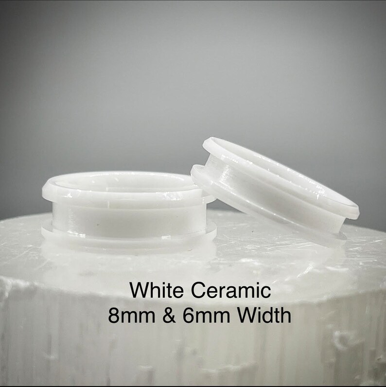 K&ouml;nnte beinhalten: Zwei Ringe aus wei&szlig;er Keramik mit glatter, gl&auml;nzender Oberfl&auml;che. Ein Ring liegt flach, w&auml;hrend der andere schr&auml;g steht. Der Text White Ceramic 8mm & 6mm Width ist unter den Ringen aufgedruckt und gibt das Material sowie die Breiten an.
