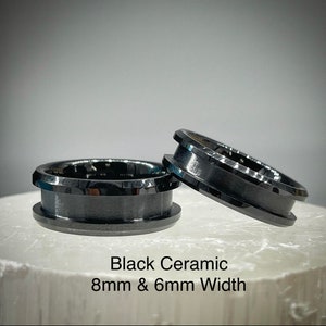 K&ouml;nnte beinhalten: Zwei schwarze Keramikringe mit polierter Oberfl&auml;che und abgeschr&auml;gten Kanten. Ein Ring liegt flach, w&auml;hrend der andere daran lehnt. Der Text Black Ceramic 8mm & 6mm Width ist unter den Ringen aufgedruckt und gibt das Material und die Breite an.