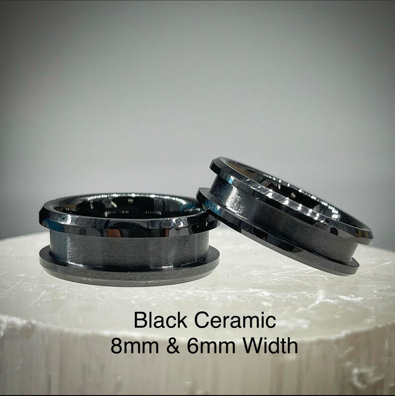 K&ouml;nnte beinhalten: Zwei schwarze Keramikringe mit polierter Oberfl&auml;che und abgeschr&auml;gten Kanten. Ein Ring liegt flach, w&auml;hrend der andere daran lehnt. Der Text Black Ceramic 8mm & 6mm Width ist unter den Ringen aufgedruckt und gibt das Material und die Breite an.