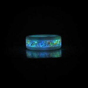 May include: A white silicone ring with a band of blue, green, and gold glitter embedded in the silicone. The ring is glowing in the dark.