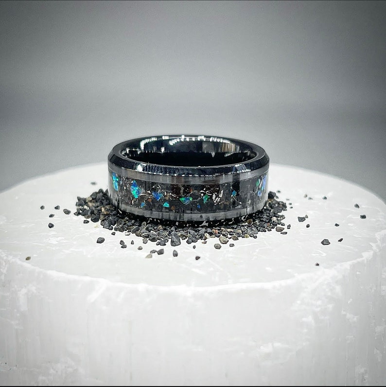 K&ouml;nnte beinhalten: Ein schwarzer Metallring mit einem breiten Band, das mit zerkleinertem dunklem Stein und schillernden blauen und gr&uuml;nen Opalfragmenten eingelegt ist. Der Ring hat eine polierte Oberfl&auml;che und ist von kleinen dunklen Steinpartikeln umgeben.