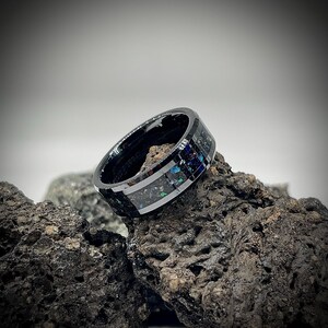 K&ouml;nnte beinhalten: Ein schwarzer Ring mit polierter Oberfl&auml;che und einer zentralen Einlage aus zerkleinertem dunklem Material mit irisierenden blauen und gr&uuml;nen Sprenkeln. Der Ring hat eine glatte, reflektierende Oberfl&auml;che und liegt auf einem dunklen, por&ouml;sen Vulkangestein.