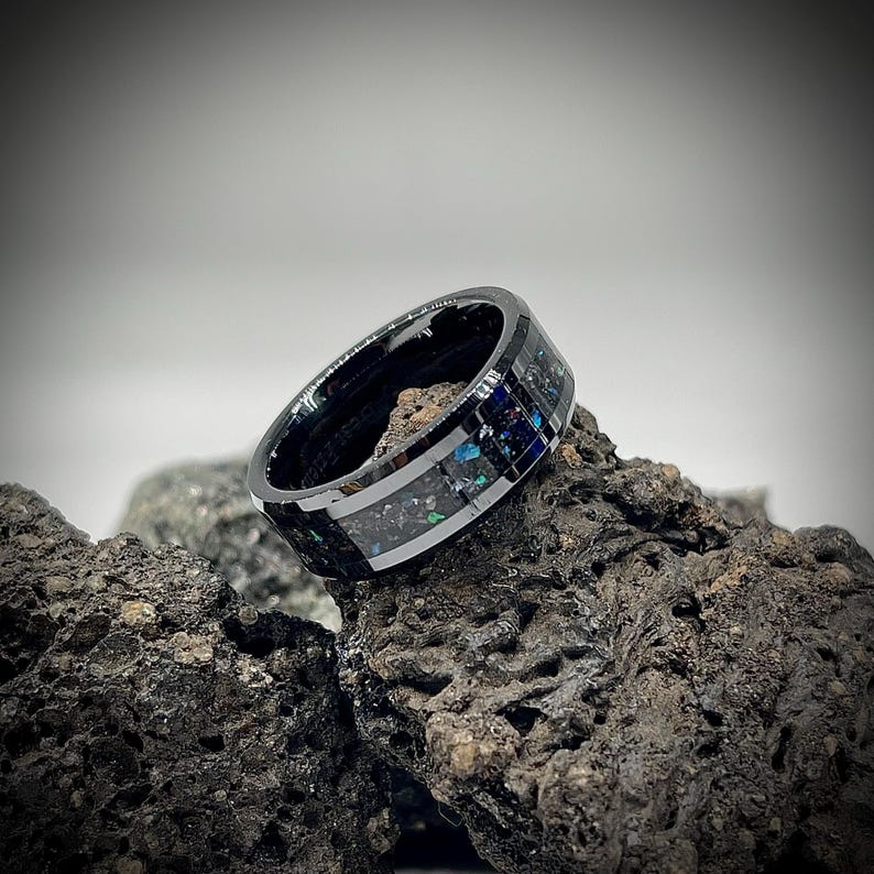 K&ouml;nnte beinhalten: Ein schwarzer Ring mit polierter Oberfl&auml;che und einer zentralen Einlage aus zerkleinertem dunklem Material mit irisierenden blauen und gr&uuml;nen Sprenkeln. Der Ring hat eine glatte, reflektierende Oberfl&auml;che und liegt auf einem dunklen, por&ouml;sen Vulkangestein.