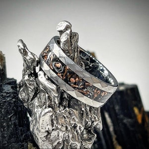 May include: A silver and black ring with a hammered finish and a copper inlay. The ring is displayed on a textured silver stand.
