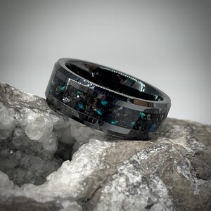 K&ouml;nnte beinhalten: Ein polierter schwarzer Ring mit einer breiten mittigen Einlage aus zerkleinertem dunklem Material und schillernden blauen sowie gr&uuml;nen Sprenkeln. Das Band hat eine glatte, reflektierende Oberfl&auml;che mit abgeschr&auml;gten Kanten.