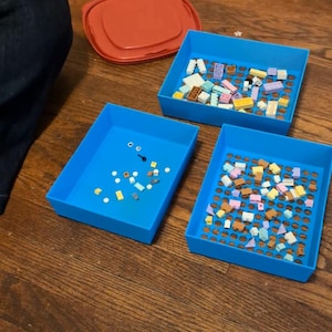 May include: Three blue plastic trays filled with colorful toy building blocks are arranged on a wooden floor. A red lid sits nearby. The blocks are various sizes and colors, including yellow, white, and brown.