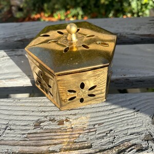 May include: A gold-coloured, hexagonal box with a lid and decorative cut-out floral patterns. The box has a small, round knob on top and is made of a shiny metal. The box is sitting on a weathered, grey wooden surface.