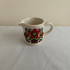 Può includere: Un piccolo contenitore per la panna in ceramica bianca con un design floreale in rosso, giallo e nero. Il design presenta un grande fiore con fiori più piccoli e foglie intorno.