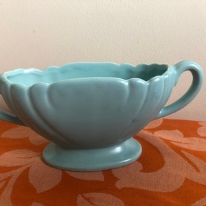 Può includere: Un vaso da fiori in ceramica blu chiaro con due manici e un bordo a festoni. Il vaso ha una base a piedistallo.